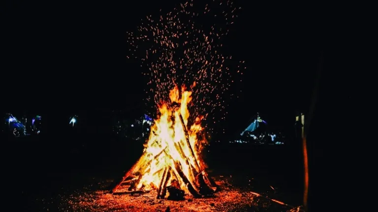 A tall bonfire of stacked logs burning at night, sending a shower of sparks into the dark sky.