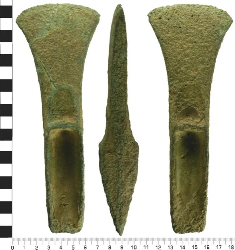 A green corroded bronze axe head photographed from three sides beside a centimetre scale, with a wide curved blade.