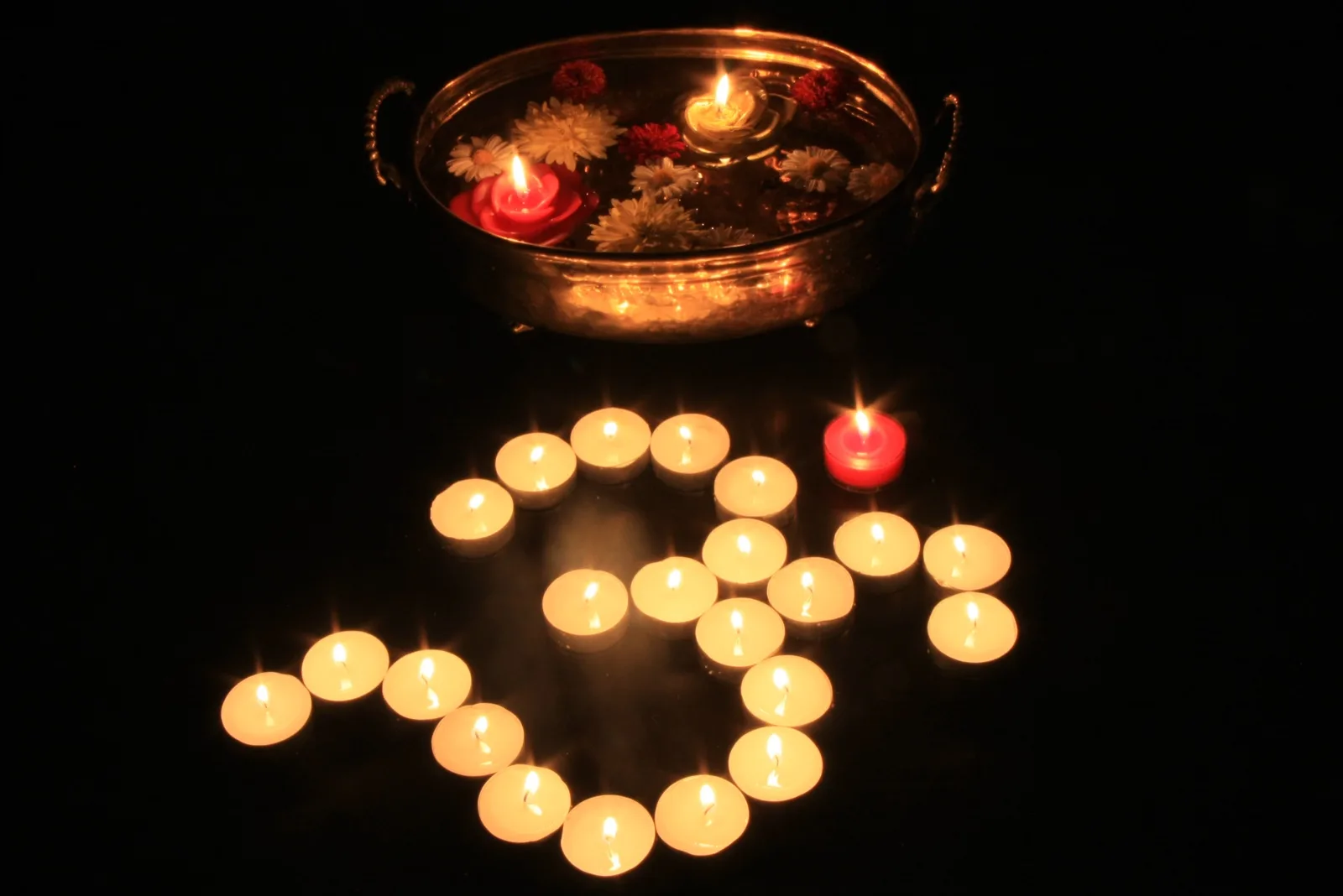 Small lit candles arranged in the shape of the Om symbol in the dark, beside a bowl of water with floating flowers and candles.