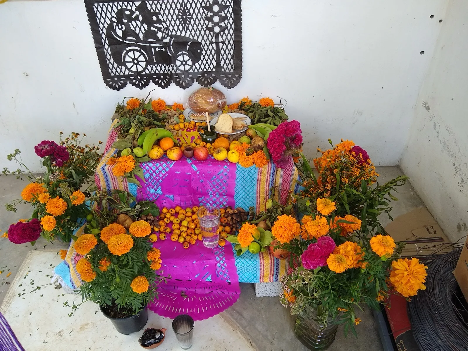 A family Day of the Dead altar in Oaxaca, Mexico, covered in orange marigolds, fruit, bread and pink cloth, with black papel picado hanging above it.