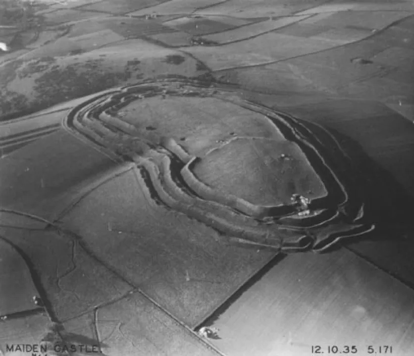 A black and white aerial photograph of a hilltop ringed by several concentric earth banks and ditches, with fields around it.