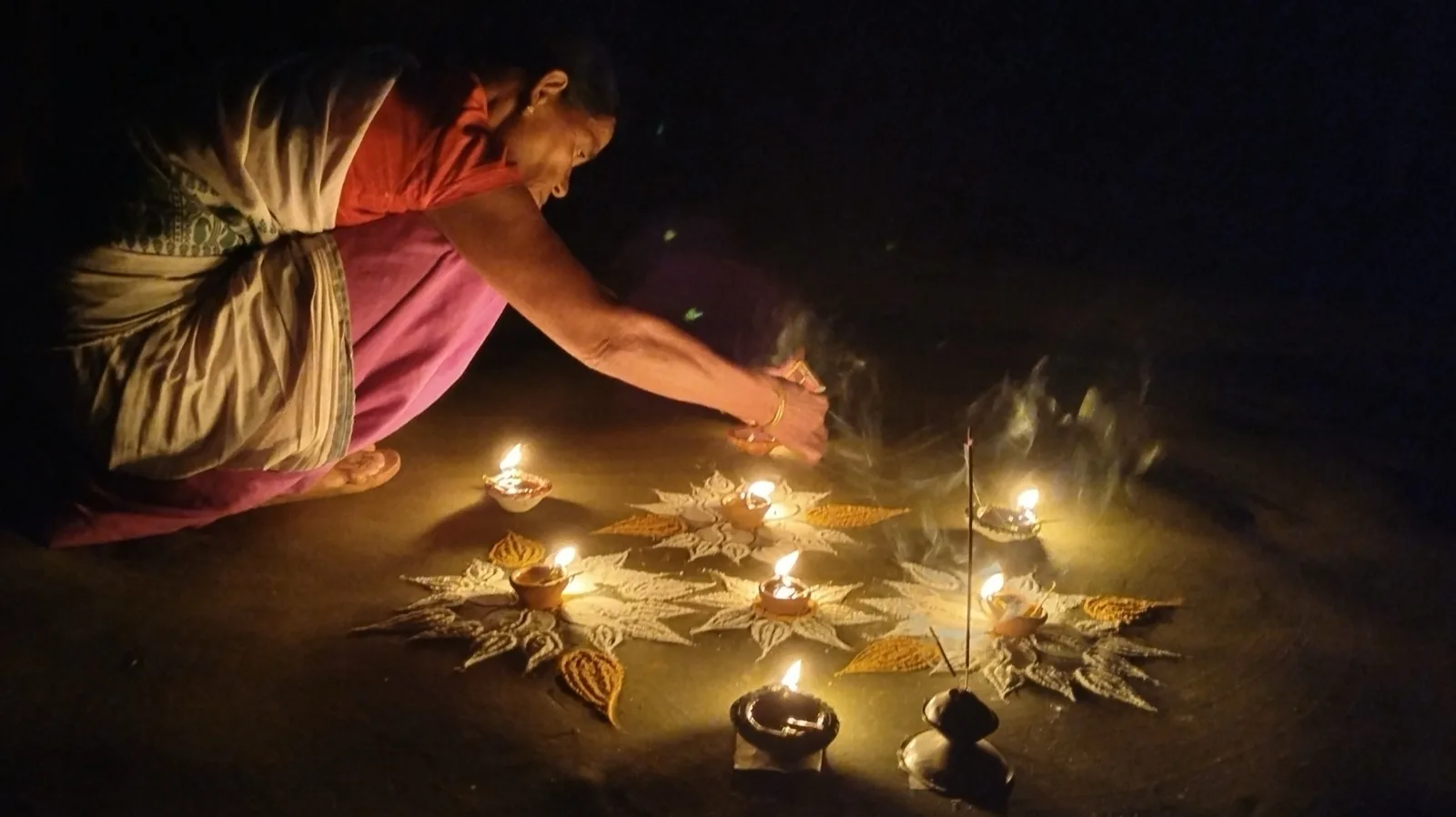 A woman bending down at night to light small clay diyas set among paper star decorations on the ground.