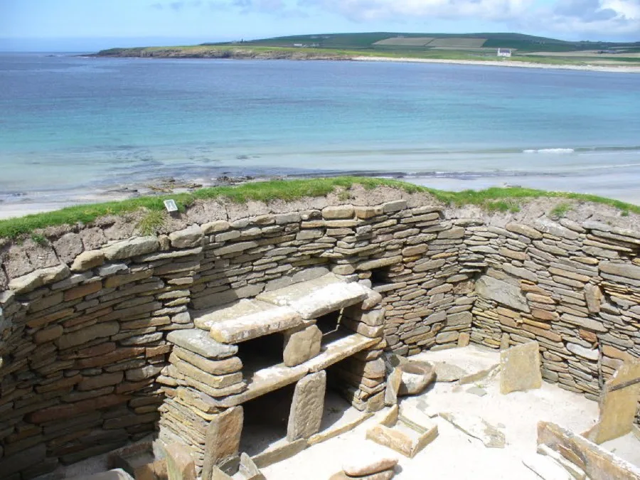 Low stone walls of a Neolithic village on a grassy shore, with a turquoise bay and headland behind.