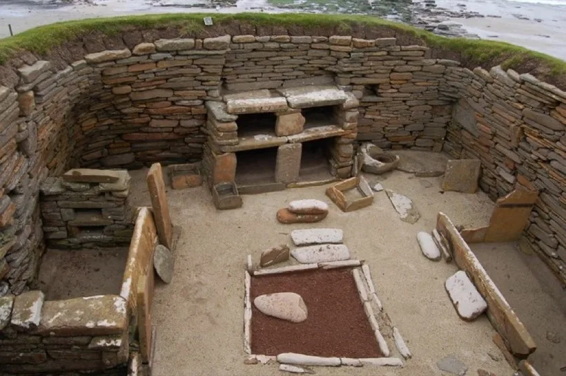 The inside of a stone-walled Neolithic house with a stone dresser against the far wall, a square hearth in the middle and stone box beds at the sides.