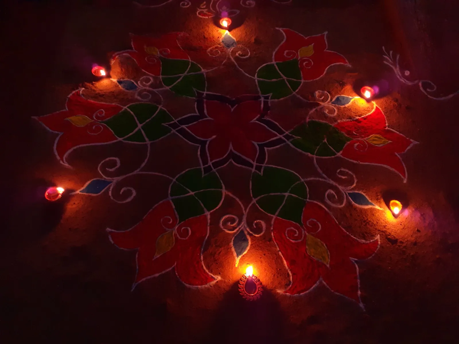 A red, green and white rangoli pattern of flowers and leaves on the ground, with lit clay diyas placed around its edge.